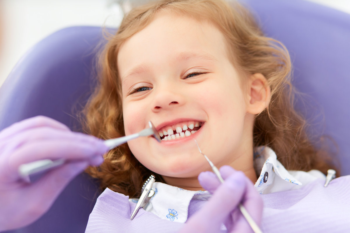 Hands of unrecognizable pediatric dentist making examination procedure for smiling cute little girl sitting on chair in hospital. Dentist office. Little girl sitting in the dentists office