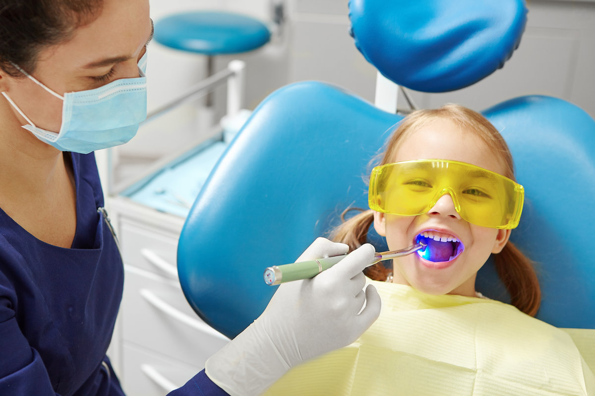 Process of drying the dental seal after treatment of the patient's tooth in pediatric dentistry. Little girl in dentist office. Doctor visiting of child.