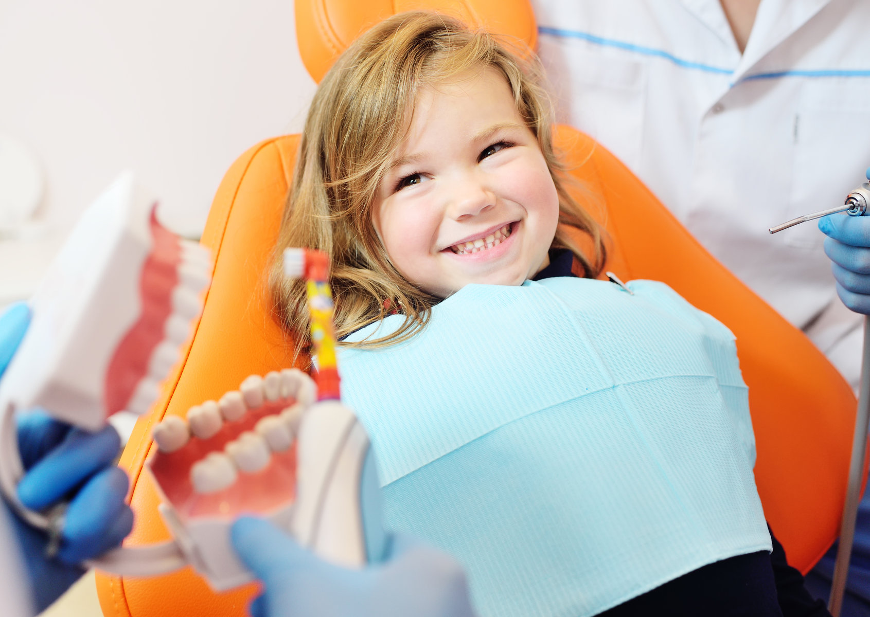 a pediatric dentist shows how to properly brush her teeth to a little girl who is sitting in an orange dental chair and smiling.